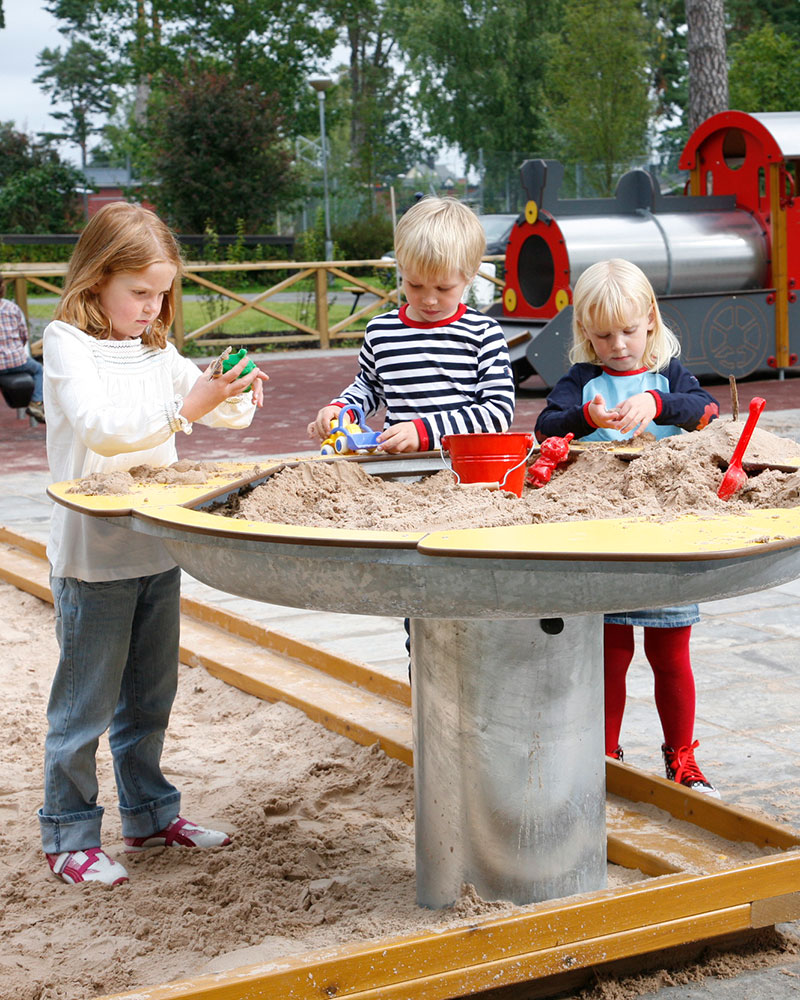 Barna leker i en sandkasse, og det er et blomsterformet sandbrett som også er fylt med sand. De kan enten stå og leke eller leke direkte i sandkassen.